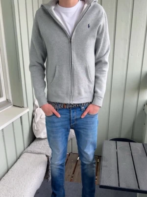 Grå hoodie från Polo Ralph Lauren - Snygg grå hoodie från Polo Ralph Lauren med dragkedja framtill och två fickor. Klassisk broderad logga på bröstet och huva. Tillverkad i mjuk bomullsmix, perfekt för chill dagar. Passar till det mesta och har en clean look.