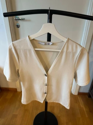 Beige v-ringad topp från 157 - Beige kortärmad topp från 157 med v-ringning och tre dekorativa knappar framtill. Modellen är croppad och har en enkel, stilren design som passar perfekt till jeans eller kjol.