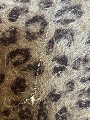 Silverhalsband med hjärta och pil litet - Snyggt halsband i silverfärg med ett glansigt hjärthänge som har en pil genom sig. Kedjan är tunn och smidig, och hänget har en cool, modern look. Perfekt accessoar för dig som gillar romantiska detaljer och vill sticka ut lite extra. Rostfri 