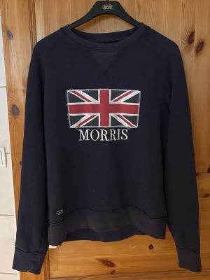 Mörkblå sweatshirt från Morris - Mörkblå sweatshirt med Union Jack-flagga och Morris-tryck på bröstet. Tröjan har rund halsringning, långa ärmar och ribbade muddar. Perfekt för en avslappnad och stilren look.