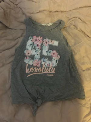 Grå linnetopp med blommor och knyt - Grå ärmlös linnetopp med stort tryck av siffran 95 dekorerad med blommor och texten 'honolulu hawaii' framtill. Toppen har knytning nedtill för en avslappnad look. Perfekt för varma dagar och somriga vibes.
