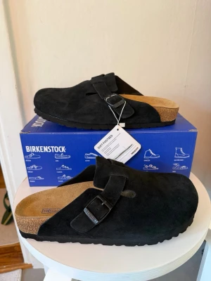 Birkenstock Boston svarta tofflor - Svarta Birkenstock Boston tofflor i mocka med klassisk design och justerbart spänne. Fotbädden är i kork och ger skön komfort. Perfekta för en chill och stilren look. Passar dig som gillar tidlös och enkel stil.