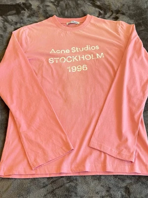 Rosa långärmad tröja Acne Studios - Snygg rosa långärmad tröja från Acne Studios med vit text 'Acne Studios STOCKHOLM 1996' framtill. Tröjan har rund halsringning och är gjord i mjuk bomull. Perfekt för en avslappnad och trendig look.
