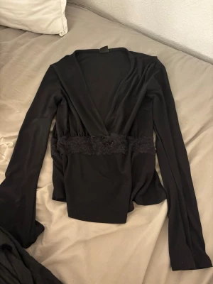 Svart omlottopp från Gina Tricot XS - Svart långärmad omlottopp från Gina Tricot i stretchigt material. Toppen har en markerad midja med spetsdetaljer och djup v-ringning. Perfekt att styla med jeans eller kjol för en trendig look.