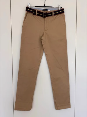 Beige chinos,  Polo Ralph Lauren, strlk 14 år - Snygga beige chinos från Polo Ralph Lauren med raka ben och klassisk passform. Byxorna har två sidofickor fram och två bakfickor med knapp. Medföljer ett randigt bälte i marinblått, rött och gult. Tillverkade i mjuk bomull för en bekväm känsla. Liten ljusare fläck på baksidan enl bild.