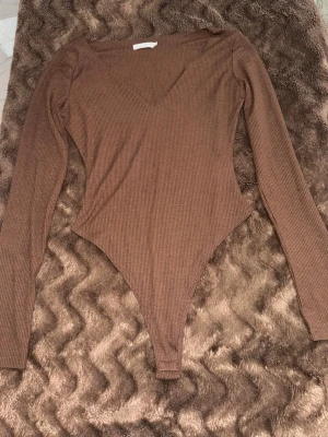 Brun ribbad body från Meshki - Säljer en snygg brun body från Meshki med lång ärm och djup v-ringning. Bodyn är ribbad och har en tight passform som framhäver figuren. Perfekt att styla med jeans eller kjol för en trendig look.