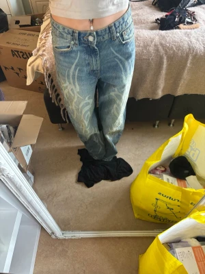 Baggy jeans med tribal-mönster - Säljer ett par ljusblå baggy jeans med vitt tribal-mönster på framsidan. Jeansen har hög midja, raka ben och klassiska fem fickor. Perfekta för en avslappnad streetstyle-look.