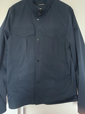 Mörkblå jacka från J.Lindeberg - Stilren mörkblå field jacket  från J.Lindeberg med tryckknappar framtill och två bröstfickor med lock. Overshirten har en rak passform, ståkrage och diskret logga på ärmen. Materialet är slätt och ger en modern look, perfekt för lager-på-lager.