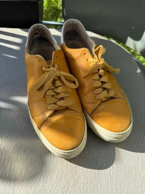 Jim Rickey Gula lädersneakers med vit sula - Snygga sneakers i gult läder med matchande gula skosnören och vit sula. Skorna har en låg profil och vit detalj på hälen. Perfekta för dig som vill sticka ut med färg och stil. Klassisk design som passar till många outfits. Lite slitna i hälen.