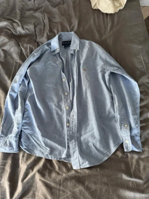 Blå skjorta från Ralph Lauren - Klassisk ljusblå skjorta från Ralph Lauren med broderad logga på bröstet. Skjortan har lång ärm, knappar framtill och är tillverkad i bomull. Perfekt för en stilren och avslappnad look.Storlek 2 vilket motsvarar ungefär xs