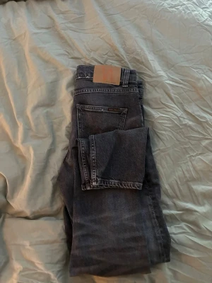 Svarta jeans från Nudie Jeans - Säljer ett par svarta jeans från Nudie Jeans med klassisk femficksmodell och snyggt läderpatch bak. Jeansen har en tvättad look och raka ben. Perfekta för dig som gillar stilrena och tidlösa plagg i garderoben. Helt nya, använda en gång. Köptes från NK för 1599.