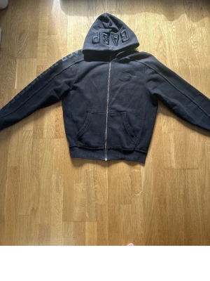 Svart GAP hoodie med dragkedja - Svart hoodie från GAP med dragkedja framtill och broderad GAP-logga på huvan. Hoodien har en stor ficka på magen och diskret broderad logga på bröstet. Tillverkad i mjukt sweatshirtmaterial och har ribbade muddar vid ärmslut och nederkant.