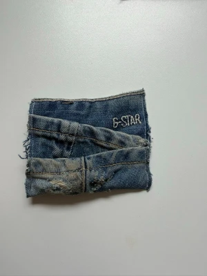 Blå jeansdetalj från G-Star - Unik jeansdetalj från G-Star i blå denim med slitna och fransiga kanter. Broderad G-Star-logga och klassisk jeanssöm ger en cool och rå känsla. Perfekt för dig som gillar streetstyle och denim med attityd.