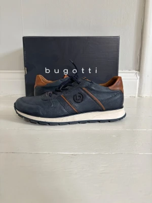 Blå sneakers från Bugatti - Snygga sneakers från Bugatti i mörkblått läder med bruna detaljer vid häl och sidor. Skorna har vita sulor, svarta snören och dragkedja på sidan för extra stil och smidighet. Perfekta för dig som vill ha en klassisk men modern look. Köpt för 1500 men säljer för 550