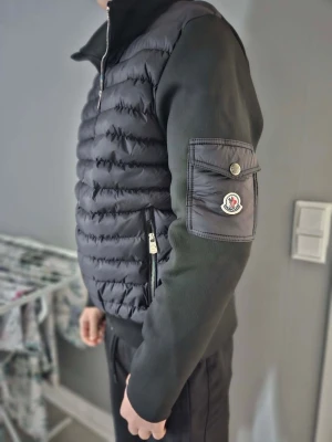 Svart pufferjacka från Moncler - Snygg svart pufferjacka från Moncler med quiltad front och släta ärmar. Jackan har dragkedja framtill, högt krage och en ficka med Moncler-logga på ärmen. Perfekt för kalla dagar och ger en stilren look.
