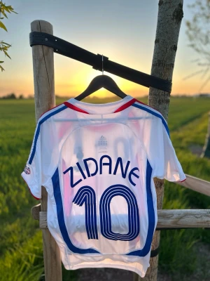 Frankrike Zidane tröja Adidas - Helt ny och oanvänd vit fotbollströja från Adidas med blå och röda detaljer, nummer 10 och Zidane på ryggen.  Frankrike-tröja med märken och tryck, kort ärm och sportig passform. Storlek M. DM för eventuella frågor eller bilder. 