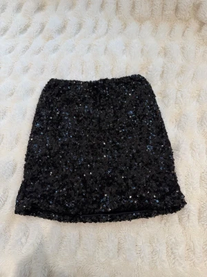 Svart paljettkjol i kort modell - Glittrig svart kortkjol täckt med paljetter som ger en snygg och festlig look. Kjolen har rak passform och är perfekt för dig som vill sticka ut. Materialet är troligtvis polyester och den är helsvart med massor av glans. Originalpris låg på 700