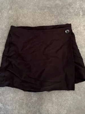 Svart skort med knappdetalj - Säljer en svart skort med omlott-look och en snygg knappdetalj framtill. Skorten har shorts under och kjol över, vilket ger en chill och trendig vibe. Perfekt för dig som vill ha både stil och bekvämlighet. Materialet känns mjukt och lätt. Ser något rosa ut på bilden men kan garantera att den är helt svart