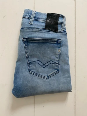 Replay Jeans  - Säljer ett par ljusblå jeans från Replay med klassisk femficksdesign och snygga detaljer på bakfickorna. Jeansen har en svart läderpatch med Replay-logga i midjan och är tillverkade i stretchig denim för en bekväm passform. Ny pris 1800kr W28/L32