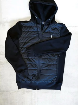 Svart cardigan från Polo Ralph Lauren - Svart cardigan från Polo Ralph Lauren med quiltad framsida, dragkedja och broderad logga på bröstet. Huva med dragsko och ribbade muddar. Perfekt för en stilren och sportig look.