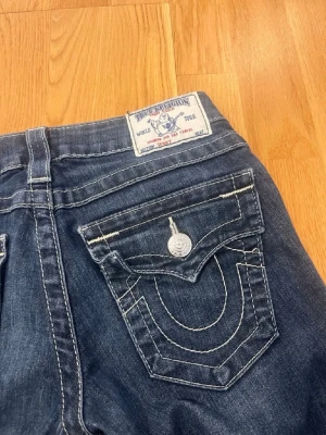 True Religion bootcut jeans - Säljer ett par bootcut mörkblå jeans från True Religion med tydliga vita kontrastsömmar och ikoniska fickdetaljer. Jeansen har klassisk femficksdesign, silverfärgade knappar och patch med logga bak i midjan. Perfekta för dig som gillar street och premium denim.