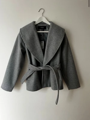 Grå kappa med skärp från Bik Bok - Stilren grå kappa från Vero moda med bred krage och knytskärp i midjan. Kappan har en rak passform och är tillverkad i ett mjukt, ull-liknande material. Perfekt för kyliga dagar när du vill vara både varm och snygg.