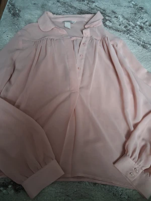 Ljusrosa blus med knappar och krage - Söt ljusrosa blus i ett tunt, lätt och transparent tyg. Blusen har klassisk krage, knäppning framtill och långa ärmar med breda manschetter och knappar. Perfekt för en chic och avslappnad look. Den har lite missfärgning på baksidan, men inget man märker av. Annars i topp-skick! 