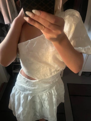 Vit asymmetrisk klänning med struktur - Säljer en vit kortklänning med ett vackert blommigt strukturmönster. Klänningen har en axelbandslös design på ena sidan och en puffärm på andra, vilket ger en unik och trendig look. Kjolen är voluminös och materialet känns lyxigt och elegant.