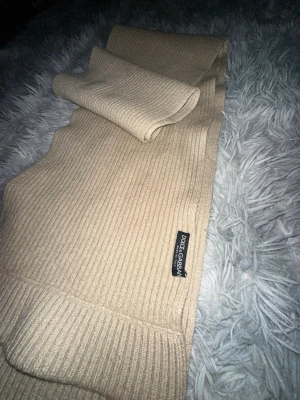 Beige halsduk från Dolce & Gabbana - Ribbstickad beige halsduk från Dolce & Gabbana. Tillverkad i Italien med klassisk design och diskret logotypmärke. Perfekt accessoar för att lyfta din outfit med en lyxig touch.