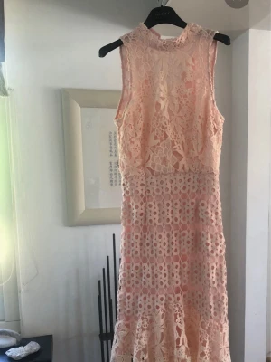 Klänning spetsklänning med blommönster - Superfin persikofärgad långklänning med blommig spets och höghalsad design. Klänningen är ärmlös och har vackra volangdetaljer nertill. Perfekt för dig som vill sticka ut med en romantisk och feminin look.