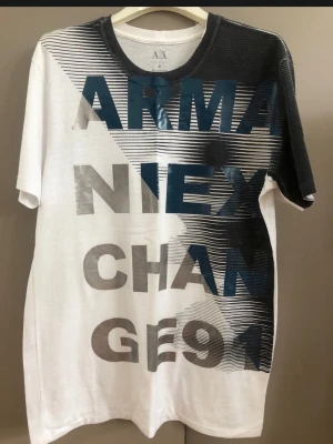 Armani Exchange grafisk t-shirt - Säljer en snygg t-shirt från Armani Exchange med stor grafisk text i blått och grått över hela framsidan. T-shirten är vit med svart och grått tryck på ena sidan och har rund hals. Perfekt för dig som gillar streetwear och vill sticka ut med en unik design.