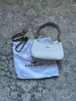 Vit axelremsväska från Coach - Snygg vit handväska från Coach med guldiga kedjedetaljer och klassiskt logomönster. Väskan har både kort axelrem och en längre avtagbar rem, samt Coach-logga i metall framtill. Tillverkad i canvas och läder. Perfekt för dig som gillar stilrena accessoarer.
