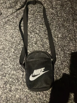 Svart axelväska från Nike - Snygg svart axelväska från Nike med vit logga framtill. Väskan har justerbar axelrem, dragkedja och röd insida med flera fack för småprylar. Perfekt för att bära med sig det viktigaste och hålla stilen.