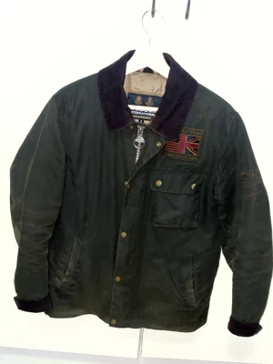 Grön Barbour jacka med patch - Klassisk grön Barbour jacka med mörkbrun krage, Union Jack-patch på bröstet och flera fickor framtill. Jackan har knappar och dragkedja samt detaljer i guld. Perfekt för dig som gillar tidlös brittisk stil.
