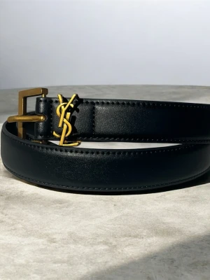 Svart YSL bälte i skinn med gulddetaljer - Stilrent svart bälte från Yves Saint Laurent  med ikoniskt YSL-monogram i guld och matchande guldfärgat spänne. Perfekt accessoar för att lyfta din outfit med en lyxig touch.