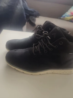 Svarta kängor med vit sula/42 - Svarta kängor med högt skaft och snörning, tillverkade i mockaliknande material. De har en vit, mönstrad sula som ger en sportig känsla. Märket verkar vara Adidas, syns på sidan av skon. Perfekta för en urban stil.