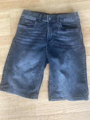 Svarta jeansshorts med fem fickor - Säljer ett par svarta jeansshorts med klassisk femficksdesign och knappgylf. Shortsen har en tvättad look och raka ben som slutar strax ovanför knäna. Perfekta för en avslappnad stil i sommar.