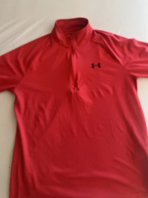 Röd träningströja från Under Armour - Sportig röd träningströja från Under Armour med halv dragkedja och kort ärm. Tröjan har en svart logga på bröstet och är tillverkad i ett lätt och snabbtorkande material, perfekt för träning och aktivitet.