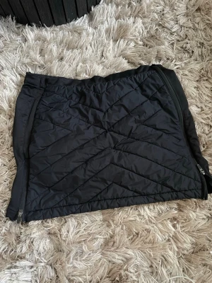Svart quiltad täck kjol från Rocktrail - Quiltad svart kjol från Rocktrail med dragkedjor på sidorna. Kjolen har en sportig look och är tillverkad i syntetmaterial, perfekt för kyligare dagar. Enkel att matcha med olika outfits och har en bekväm passform.