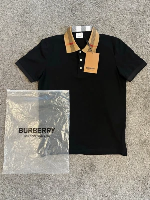 Svart Burberry pikétröja med rutig krage - Snygg svart pikétröja från Burberry med klassisk rutig krage i beige, svart och rött. Tröjan har knappar framtill och är kortärmad. Perfekt för dig som vill ha en stilren och exklusiv look.