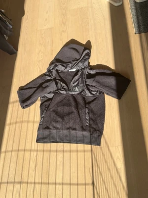 Svart jacka/hoodie från Calvin Klein - Snygg svart jacka/hoodie från Calvin Klein med dragkedja vid halsen och huva. Tillverkad i mjukt material med detaljer i blankt tyg på axlar och bröst. Stor magficka framtill och CK-logga broderad på bröstet. Perfekt för en chill och trendig look. Inte för varm så perfekt för sommar kvällar. 