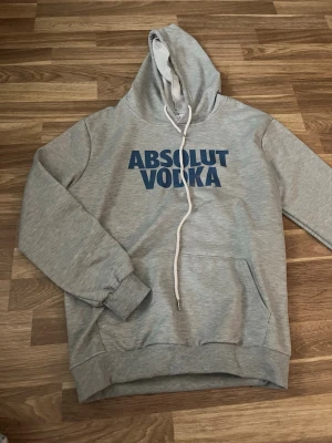 Grå hoodie med Absolut Vodka-tryck - Snygg grå hoodie med blått 'ABSOLUT VODKA'-tryck på bröstet. Klassisk modell med huva och vita dragsnören samt känguruficka framtill. Tillverkad i mjukt bomullsmaterial, perfekt för en avslappnad streetstyle.