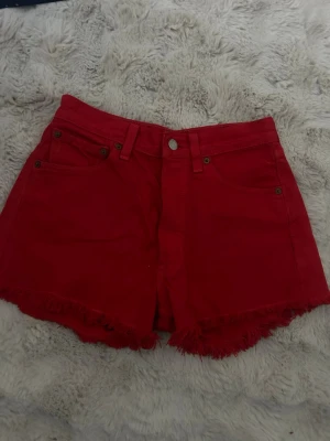 Röda Levi's 501 shorts med fransar - Snygga röda Levi's 501 shorts i klassisk femficksmodell. Shortsen har råa fransade benslut, knappgylf och Levi's ikoniska läderpatch bak i midjan. Perfekta för sommaren och ger en cool vintagekänsla.
