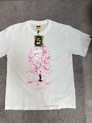 Vit BAPE t-shirt med rosa tryck - Säljer en clean vit t-shirt från BAPE med ett stort rosa tryck av ett träd och BAPE-loggan på bröstet. Klassisk passform och rund hals. Ikonisk BAPE-tag på ärmen och originaltag kvar. Perfekt för dig som gillar streetwear och vill sticka ut.