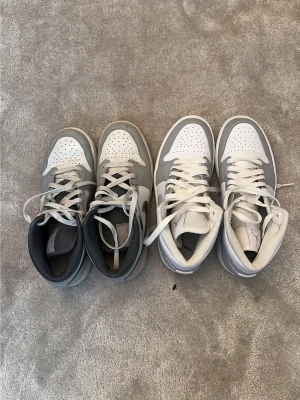Nike Air Jordan 1 Mid Grå/Vit - Säljer två par Nike Air Jordan 1 Mid sneakers i grått och vitt. Klassisk siluett med perforerad tå, vita skosnören och ikoniska swoosh-loggan på sidan. Skorna har en mix av läder och syntetmaterial för en clean och sportig look. Dom blå vita skorna är helt nya och dom gråsvarta är lite använda. Blå/vita skorna: strl 38,5, svart grå skorna: strlk 39