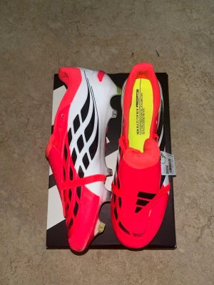 Adidas Predator PRO  - Säljer ett par Adidas Predator fotbollsskor i en riktigt fet rosa och vit färg med svarta detaljer. Skorna har låg profil, snörning och mönstrad sula för bra grepp på planen. Ovandelen är i syntetmaterial och designen är modern och sportig.