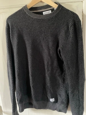 Mörkgrå stickad tröja Jack & Jones - Snygg mörkgrå stickad tröja från Jack & Jones med diskret strukturmönster. Tröjan har rund halsringning och ribbade muddar vid ärmslut och nederkant. Perfekt för lager på lager och enkel att matcha med jeans eller chinos.