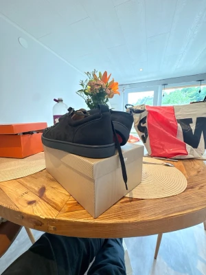 Svarta Louboutin sneakers med nitar - Säljer ett par svarta sneakers från Christian Louboutin med mattsvarta nitar på tån och klassisk röd sula. Skorna har lågt skaft, svarta skosnören och är tillverkade i mocka och läder. Perfekta för dig som vill sticka ut med en edgy vibe. (Storlek 45)