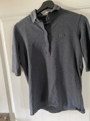 Mörkgrå pikétröja från Lacoste - Mörkgrå pikétröja från Lacoste med klassisk krage och knappslå framtill. Tröjan har korta ärmar och den ikoniska krokodilloggan broderad på bröstet. Perfekt för en stilren och avslappnad look.