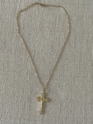Guldfärgat halsband med korshänge - Snyggt guldfärgat halsband med ett detaljerat korshänge. Korset har små färgade stenar i blått, orange och vitt samt en Jesusfigur i mitten. Kedjan är tunn och elegant, perfekt för dig som gillar klassiska smycken med en twist.Läng: 40+6cm Kors: höjd 4,2cm, Breda: 2,5cm